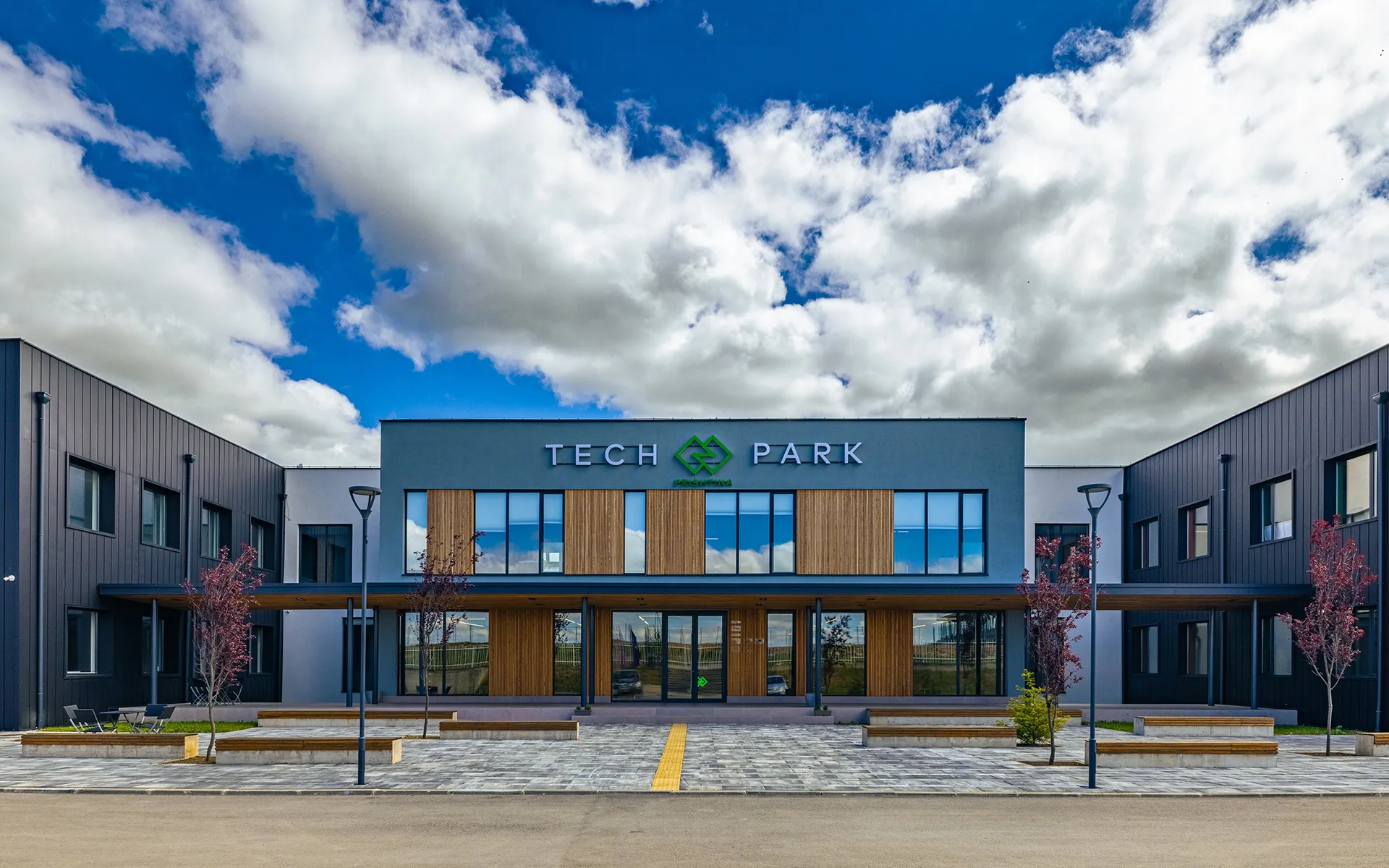 The Tech Park Prishtina building: a charcoal-clad facade with warm wood panels and a dramatic clouded sky, fronted by a paved plaza with young trees.